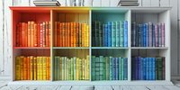 Books arranged by color in rainbow spectrum order on white wooden shelves, creating vibrant gradient display from red through orange, yellow, green to blue tones.