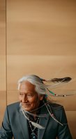 Senior man with long white and dark hair adorned with colorful beads and feathers, wearing dark blazer and turquoise jewelry against neutral beige wall background.