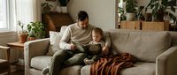 Father and toddler son bonding on living room sofa, reading book together in cozy home interior with plants and natural light for family lifestyle content.