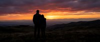 Couple silhouettes embracing while watching dramatic sunset over mountain landscape with orange and purple sky colors creating romantic outdoor moment.