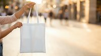 Woman holding white cotton tote bag on urban street with blurred shopping district background, suitable for retail, eco-friendly shopping, branding mockups and sustainable lifestyle marketing.