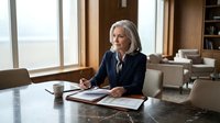 Senior woman with white hair, Caucasian ethnicity, wearing dark blazer and white blouse, sitting at modern desk with laptop and documents in contemporary office with large windows and bookshelves.