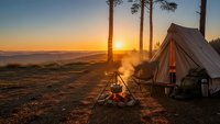 Camping tent and campfire at golden sunset with mountain views. Outdoor adventure and wilderness recreation scene for travel and nature enthusiasts.