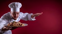 Caucasian male chef in white uniform and hat presenting gourmet pasta dish with dramatic gesture against dark red background, professional restaurant service.