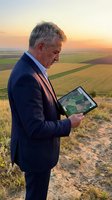 Senior businessman using tablet for agricultural planning at sunset overlooking farmland fields for precision farming and crop management technology.