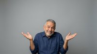 Middle-aged man with gray beard and dark hair, wearing navy blue shirt, gesturing with both palms up in confused or uncertain expression against neutral gray background.