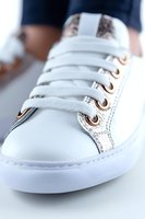 Close-up view of white leather sneaker with metallic rose gold eyelets and white laces against blurred background, showcasing modern casual footwear style.