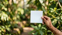 Female hand with henna tattoo holding blank white card against lush green garden foliage background for mockup and branding purposes.