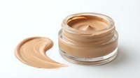 Open glass jar of beige foundation makeup with creamy texture and smooth swirl sample on white background for cosmetic and beauty product advertising.