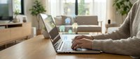 Woman working on laptop at wooden desk in bright modern living room with cozy beige sweater for remote work and home office productivity concepts.