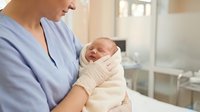Nurse in blue scrubs gently holds a swaddled newborn baby in a hospital maternity ward, conveying care and new life.