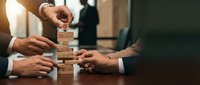 Business team building wooden block tower together demonstrating teamwork collaboration strategy planning and problem solving skills in office meeting.
