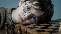 Young boy with glasses resting head on chess board during strategic game thinking about next move in intellectual competition.