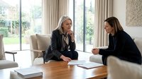 Two professional businesswomen having serious discussion at modern office table reviewing documents and contracts in bright contemporary workspace.
