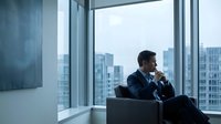 Thoughtful businessman in suit sitting by office window overlooking city skyline, contemplating business decisions and future planning opportunities.