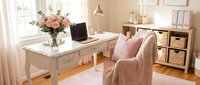 Bright home office workspace with white desk, laptop, fresh flowers in vase, pink upholstered chair, wooden storage shelves, and natural window light for professional work environment.