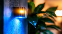 Modern outdoor wall light fixture emitting bright blue illumination against dark wall, with blurred green plant leaves in foreground creating atmospheric evening ambiance.