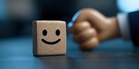 Wooden cube with happy smiley face emoji on dark background with blurred hand, representing positive attitude and customer satisfaction in business concept.