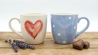 Pair of ceramic mugs on wooden surface with lavender sprigs and chocolate hearts, one cream mug featuring red heart design, one blue mug with polka dot pattern, perfect for Valentine's Day, anniversaries, or romantic gift sets.