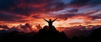 Person celebrating success on mountain peak at dramatic sunset with arms raised against colorful sky and mountain range silhouette for motivation concepts.