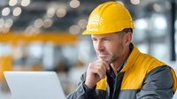Construction worker in yellow hard hat and safety vest using laptop computer at industrial site with thoughtful expression and blurred warehouse background.