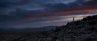 Lone hiker silhouette standing on rocky mountain ridge during dramatic sunset with purple and orange clouds over desert landscape wilderness adventure.