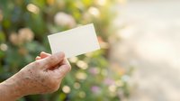 Senior person holding blank white card outdoors with blurred garden background, suitable for business cards, invitations, announcements, and promotional messaging.