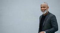 Mature African American male professional wearing dark blazer and black shirt, smiling confidently against neutral gray background. Corporate portrait for business, leadership, and executive use.