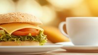 Fresh cheeseburger with lettuce, tomato, and cheese on sesame bun served on white plate with coffee cup in soft focus background. Food photography.