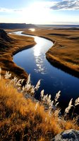 Meandering river through golden grasslands at sunset, with sunlight reflecting off water surface amid dramatic cliffs and swaying reeds in foreground.