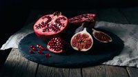 Fresh pomegranate and figs sliced on dark slate board, moody food styling with rich red tones on rustic wooden table.