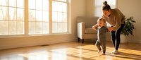 Young mother helping toddler take first steps in bright living room with natural sunlight streaming through large windows creating warm atmosphere.