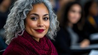 Confident mature woman with natural gray curly hair and red lipstick wearing burgundy scarf smiles warmly against blurred background. Professional portrait.