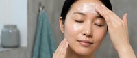 Young Asian woman applying white facial cream moisturizer in modern bathroom during morning skincare routine with eyes closed and peaceful expression.