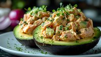 Stuffed avocado halves filled with creamy chicken salad topped with fresh microgreens on white plate, healthy lunch meal ready to eat.