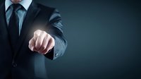 Businessman in dark suit pointing finger forward against black background, demonstrating leadership and decision-making in corporate environment.