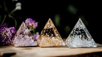 Crystal pyramids with delicate floral inclusions in pink, gold and clear resin catch light on wooden surface against dark background with purple flowers.