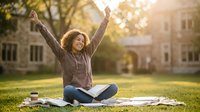 Young African American woman celebrating success while studying outdoors on campus lawn with books and coffee cup during golden hour sunset.