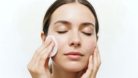 Young woman applying facial cleanser with cotton pad during skincare routine, eyes closed, gentle cleansing motion for healthy glowing skin care regimen.
