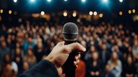 Close-up view of hand holding microphone against blurred crowd background with stage lights, capturing live performance atmosphere at concert or event venue.