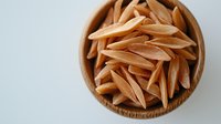 Uncooked whole grain penne pasta in wooden bowl on white background, top view close up macro shot of raw organic brown rice pasta noodles.