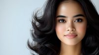 Young Asian woman with natural makeup and healthy glowing skin against light background. Dark wavy hair frames gentle smile and confident expression.