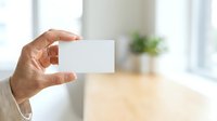 Man holding blank white business card in modern office environment with natural light and blurred background, suitable for corporate branding and professional networking concepts.
