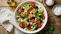 Fresh pasta salad with grilled salmon, cherry tomatoes, cucumber and basil in white bowl on rustic wooden table with olive oil and seasonings for healthy meal.