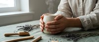 Woman holding white ceramic mug with steam rising, sitting at wooden table with dried lavender flowers scattered around, cozy morning atmosphere indoors.