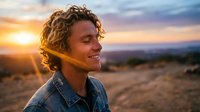 Young man with curly hair smiling peacefully with eyes closed during golden hour sunset in desert landscape, expressing joy and freedom outdoors.