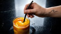 Person holding black straw in glass of golden orange juice or beverage on dark wet surface, suitable for food and drink marketing.