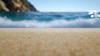 Sandy beach close-up with blurred blue ocean waves and rocky cliff in background, creating serene coastal landscape for travel and vacation concepts.