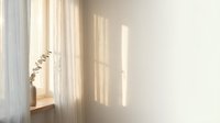 Soft morning sunlight streaming through white curtains creating gentle shadows on minimalist interior wall with dried eucalyptus branch for peaceful home decor.