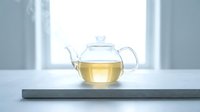 Glass teapot filled with golden herbal tea on gray slate coaster by bright window, suitable for beverage, wellness and lifestyle design projects.